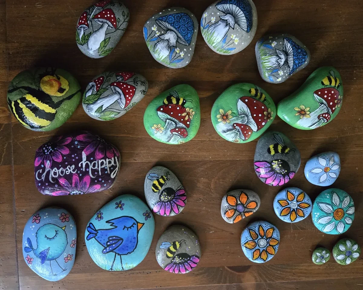 A table covered in Susan's painted stones: bumblebees, bluebirds, red-capped toadstools, flowers and a purple 'choose happy' stone